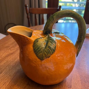 May include: A ceramic pitcher shaped like a round orange fruit with a glossy orange finish, a textured surface, and a green leaf detail at the base of the curved green handle. The pitcher features a wide pouring spout and a smooth interior.