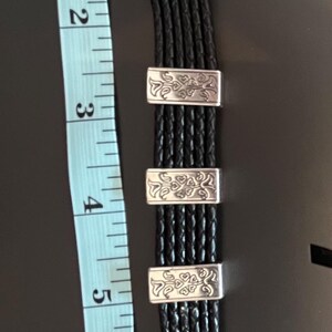 Op de afbeelding: Zwart leren armband met zilverkleurige details. De armband heeft meerdere gevlochten strengen en decoratieve rechthoekige sluitingen. De armband is ongeveer 18 cm lang.