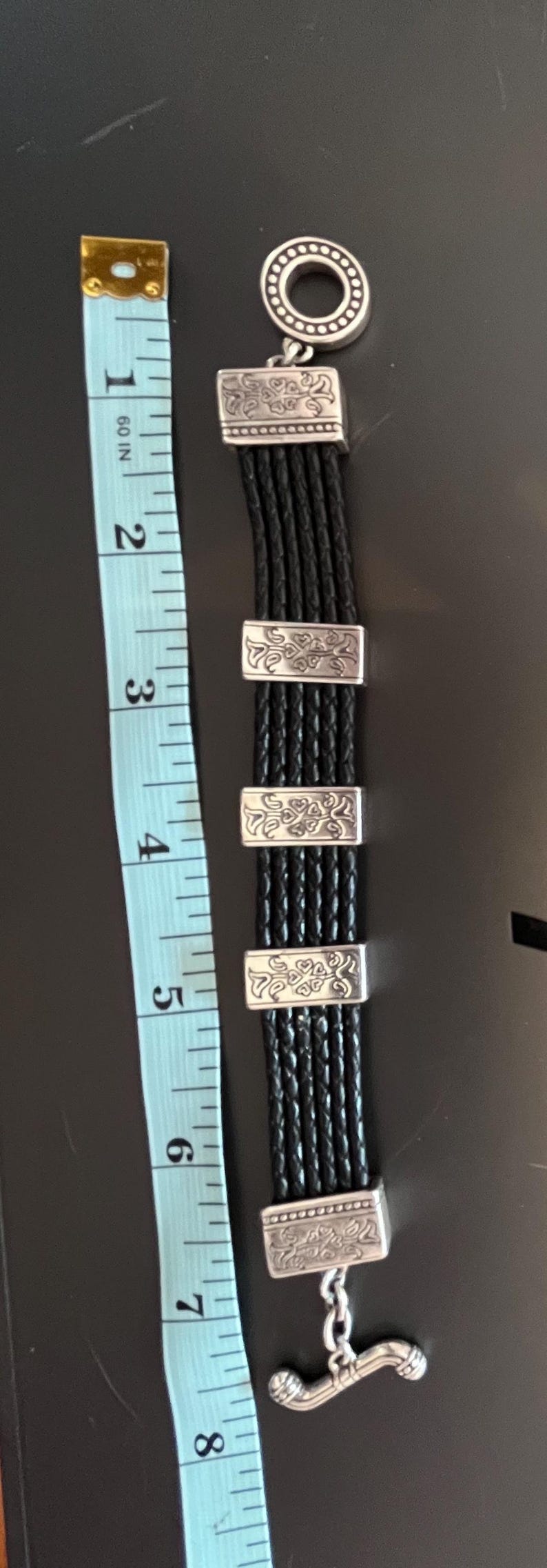 Op de afbeelding: Zwart leren armband met zilverkleurige details. De armband heeft meerdere gevlochten strengen en decoratieve rechthoekige sluitingen. De armband is ongeveer 18 cm lang.