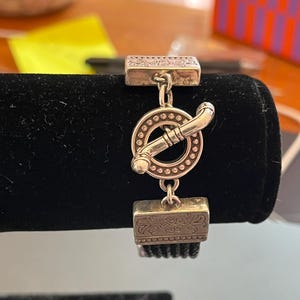 Op de afbeelding: Zilverkleurige armband met een tuimelsluiting. De armband heeft rechthoekige decoratieve elementen en een cirkelvormige sluiting met een staaf. De armband wordt gepresenteerd op een zwart fluwelen oppervlak.