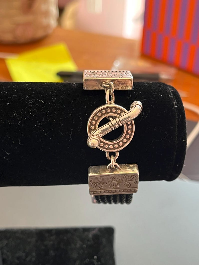 Op de afbeelding: Zilverkleurige armband met een tuimelsluiting. De armband heeft rechthoekige decoratieve elementen en een cirkelvormige sluiting met een staaf. De armband wordt gepresenteerd op een zwart fluwelen oppervlak.