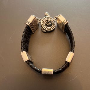 Op de afbeelding: Zwart gevlochten leren armband met zilverkleurige metalen accenten. De armband heeft vierkante en rechthoekige metalen stukken en een ronde sluiting met een Keltisch knoopontwerp. Ontworpen voor een pols.