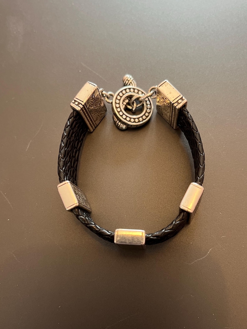 Op de afbeelding: Zwart gevlochten leren armband met zilverkleurige metalen accenten. De armband heeft vierkante en rechthoekige metalen stukken en een ronde sluiting met een Keltisch knoopontwerp. Ontworpen voor een pols.