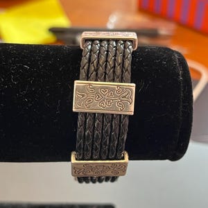 Op de afbeelding: Zwart gevlochten leren armband met zilverkleurige metalen accenten. De armband heeft geweven leren strengen die worden samengehouden door rechthoekige sluitingen met decoratieve bloempatronen. De armband wordt gepresenteerd op een zwarte fluwelen ondergrond.