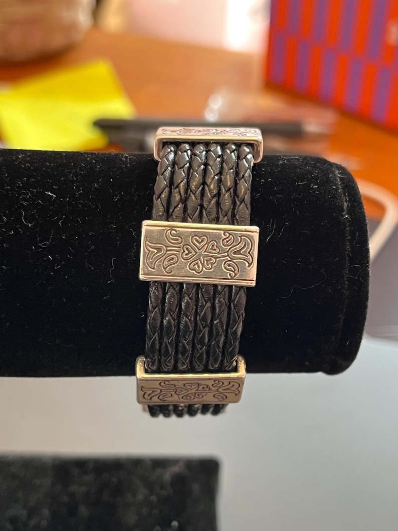 Op de afbeelding: Zwart gevlochten leren armband met zilverkleurige metalen accenten. De armband heeft geweven leren strengen die worden samengehouden door rechthoekige sluitingen met decoratieve bloempatronen. De armband wordt gepresenteerd op een zwarte fluwelen ondergrond.
