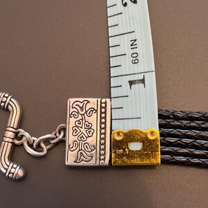 Op de afbeelding: Close-up van een zwart leren armband met een zilverkleurige sluiting en decoratieve accenten. De armband wordt gemeten met een blauwe meetlint, met een breedte van ongeveer 2,5 cm.