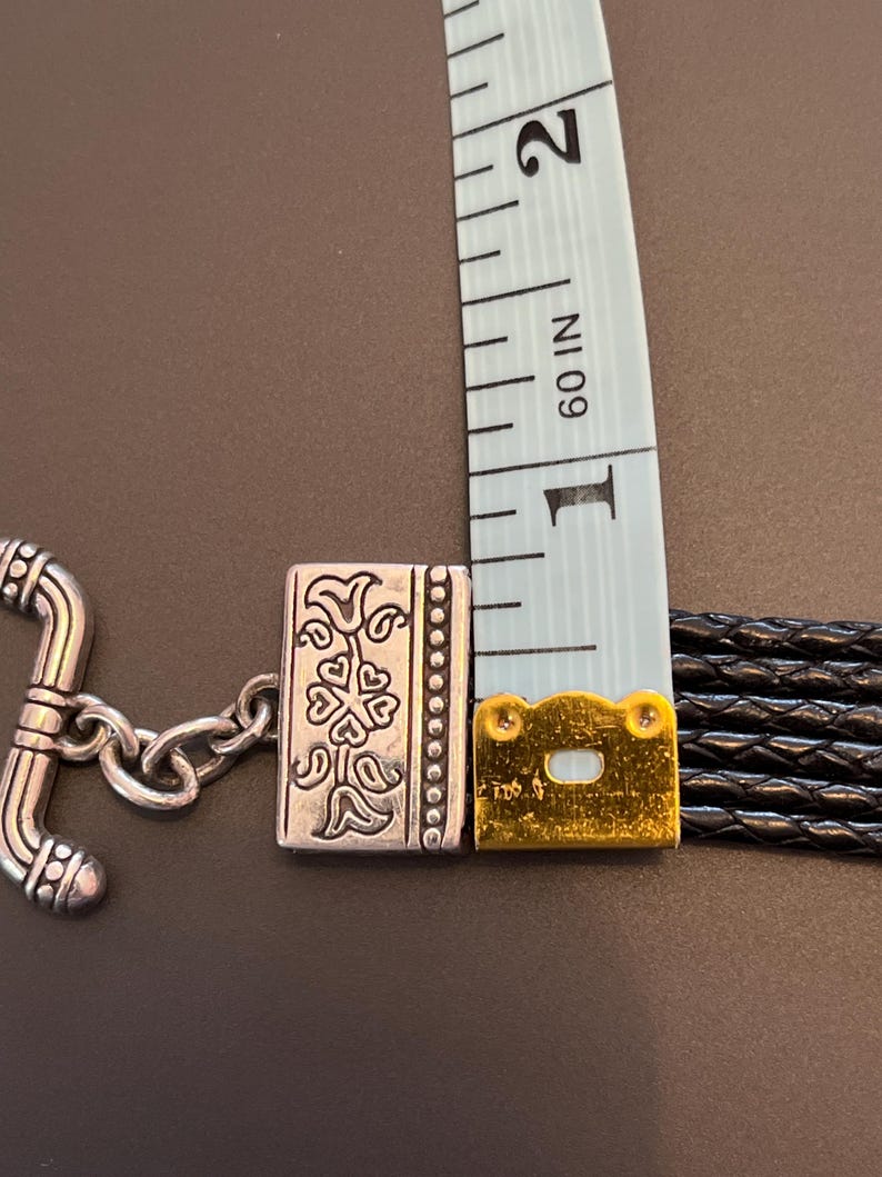 Op de afbeelding: Close-up van een zwart leren armband met een zilverkleurige sluiting en decoratieve accenten. De armband wordt gemeten met een blauwe meetlint, met een breedte van ongeveer 2,5 cm.