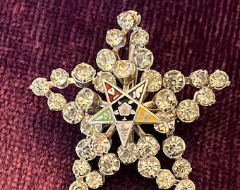 May include: A silver five-pointed star brooch with a colorful five-pointed star in the center. The brooch is decorated with clear rhinestones.