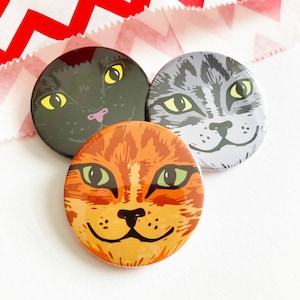 May include: Three round cat face buttons. The first button is black with a white cat face. The second button is grey with a white cat face. The third button is orange with a white cat face.
