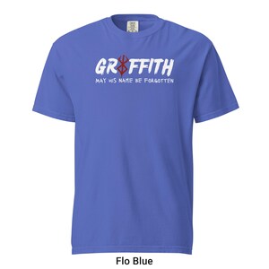 May include: A blue t-shirt with white text that reads "Griffith May His Name Be Forgotten" and a red symbol.
