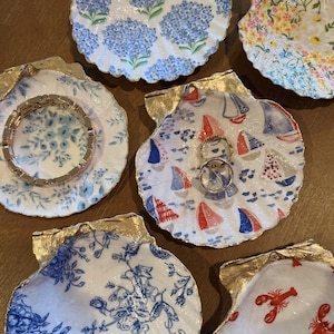 Puede incluir: Seis platos decorativos para baratijas con forma de concha, con detalles dorados. Cada plato presenta un diseño diferente en azul y blanco, incluyendo motivos florales, náuticos y de langosta.