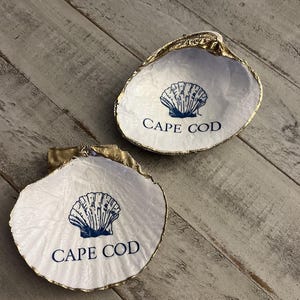 以下が含まれることがあります： 金色の縁取りが施された2つの白い貝殻の皿。各皿には青い貝殻のグラフィックと、青で印刷された「CAPE COD」の文字があります。皿は、風化した灰色の木製の表面に表示されています。
