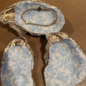 May include: Three oyster shells with a gold rim and a blue and white damask pattern. The top shell has a gold chain bracelet inside.