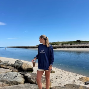 May include: A navy blue sweatshirt with the text "what the shell" and a seashell graphic. The person is standing on a beach with a blue sky and water in the background. They are holding a beverage. The scene is set on a sunny day.