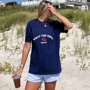 以下が含まれることがあります： 「WHAT THE SHELL」と「Cape Cod」の文字が白でプリントされたネイビーブルーのTシャツ。赤い貝殻のグラフィックがあります。人物は水色のショートパンツを着用し、飲み物を持っています。