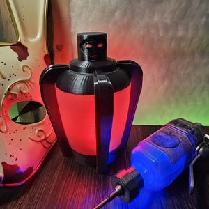 Puede incluir: Una lámpara decorativa negra y roja con un diseño único, con un interior rojo brillante. Un objeto azul y negro con forma de jeringa está al lado de la lámpara. Una máscara blanca y roja también está en la imagen.
