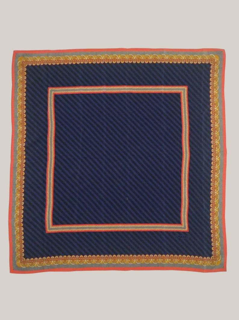 May include: Square navy blue scarf with diagonal stripes. The scarf has a red border with a gold scalloped design. A smaller red and gold square border frames the navy blue center.
