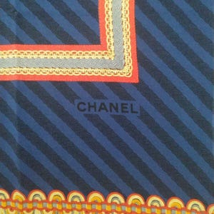 May include: A blue scarf with diagonal navy stripes and decorative borders. The borders feature red, gray, and yellow accents. The word "CHANEL" is printed in black on the scarf.