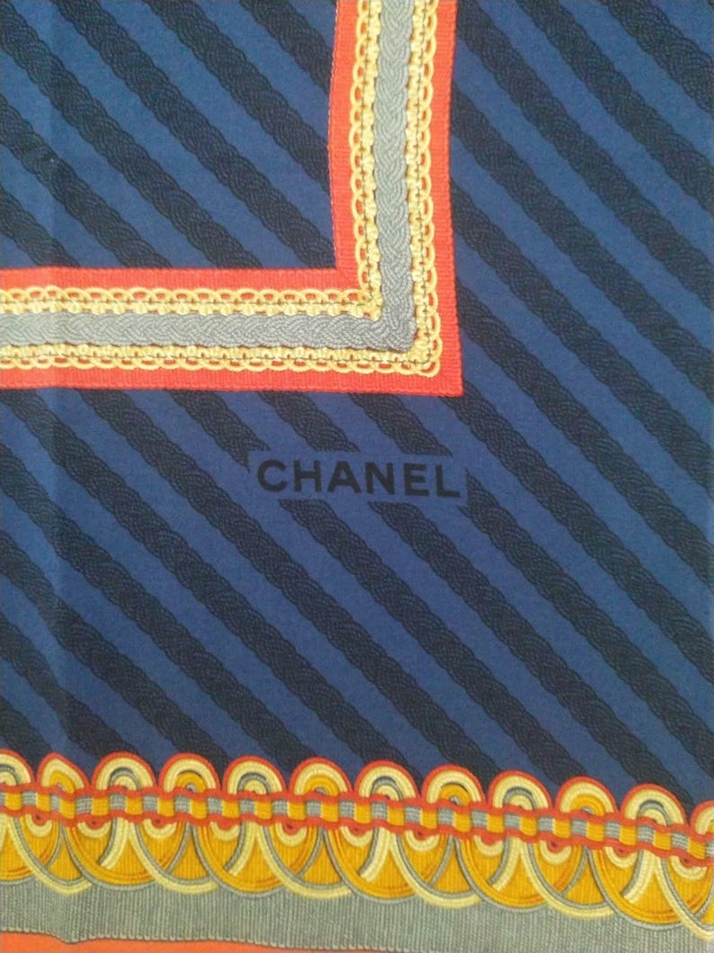 May include: A blue scarf with diagonal navy stripes and decorative borders. The borders feature red, gray, and yellow accents. The word "CHANEL" is printed in black on the scarf.