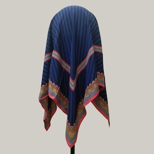 May include: A navy blue scarf with vertical stripes and decorative borders. The scarf has a repeating pattern of red, yellow, and blue accents along the edges. The scarf is draped over a stand, showcasing its design.