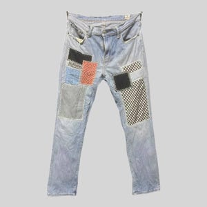 May include: Light blue denim jeans featuring a variety of rectangular fabric patches sewn onto the front. The patches include solid black, orange textured, blue and white striped, and black and white checkered patterns with visible white stitching.