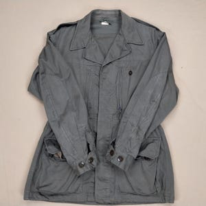 May include: A gray military-style jacket with a collared neckline, buttoned cuffs, and multiple pockets. The jacket has a zipper closure and a tailored fit. The fabric appears to be a durable, possibly cotton blend. The jacket is laid flat on a neutral background.