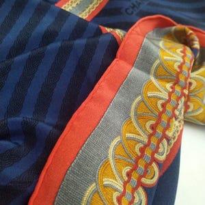 May include: A folded navy blue silk scarf with a striped pattern and decorative border. The border features a gray background with yellow and red circular designs. The scarf has a red border and the word "CHA" is visible.