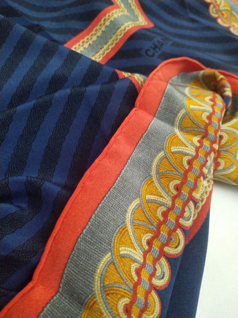 May include: A folded navy blue silk scarf with a striped pattern and decorative border. The border features a gray background with yellow and red circular designs. The scarf has a red border and the word "CHA" is visible.