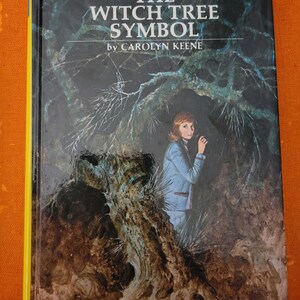 Op de afbeelding: De omslag van een Nancy Drew-mysterieboek getiteld "The Witch Tree Symbol" door Carolyn Keene. De omslag toont een jonge vrouw die in een donker bos staat en over haar schouder kijkt. Het boek is nummer 33 in de serie.