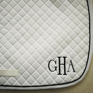 May include: White quilted saddle pad with black trim and the embroidered initials "GHA" in black. The pad has a diamond pattern and a small white strap. Equestrian themed.