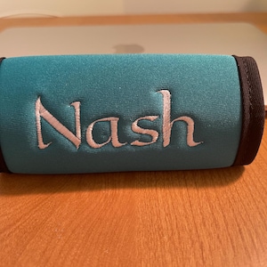 May include: Teal-colored, cylindrical item with the name "Nash" embroidered in a light pink script. The ends are trimmed with black fabric. The item is resting on a wooden surface.