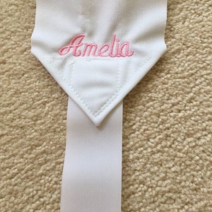 Puede incluir: Pulsera de tela blanca con el nombre "Amelia" bordado en rosa en la parte superior.
