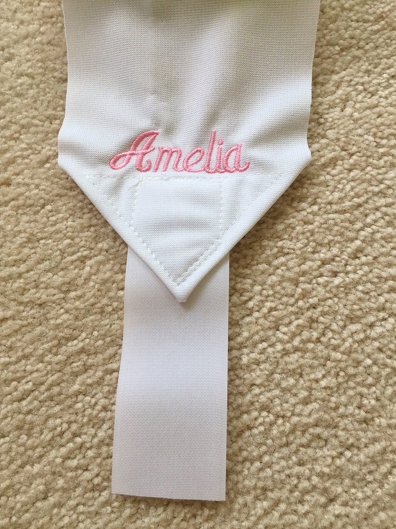 Puede incluir: Pulsera de tela blanca con el nombre "Amelia" bordado en rosa en la parte superior.