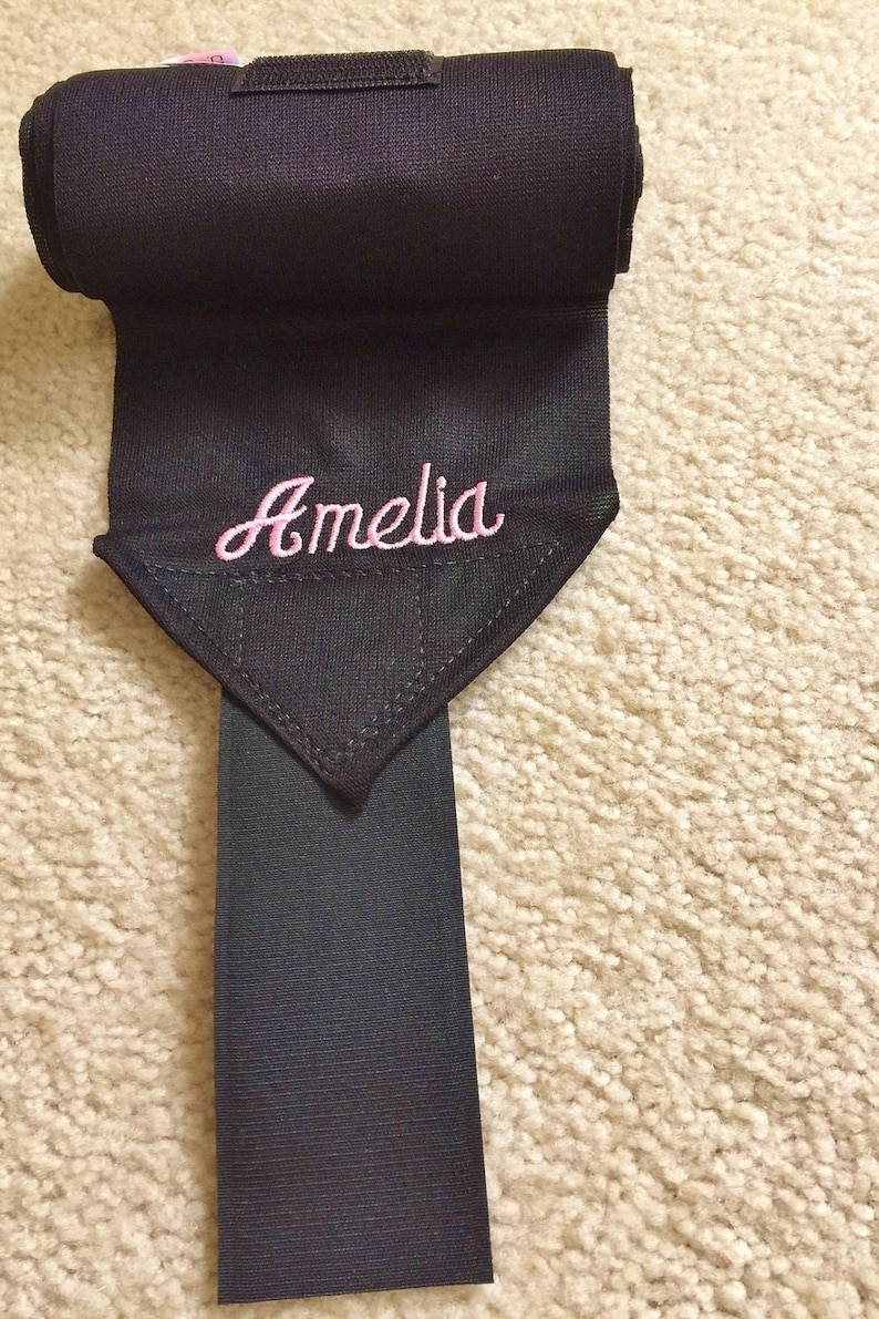 Puede incluir: Vendas negras para las piernas de caballo con un nombre bordado en rosa "Amelia" en una solapa triangular.