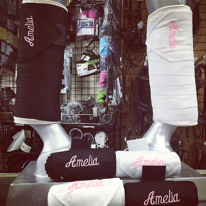 Puede incluir: Cuatro vendas para las piernas de caballo negras y blancas con bordado rosa que dice "Amelia".