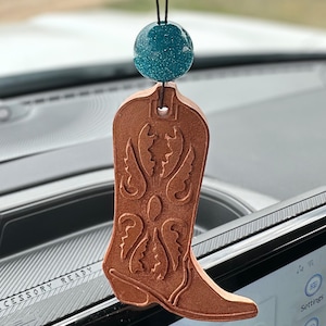 May include: A brown, western-style car air freshener shaped like a cowboy boot. The boot has decorative embossed details and hangs from a black string attached to a teal, glittery ball. The accessory is hanging in a car.