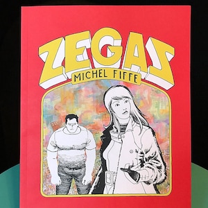 May include: A red comic book titled "ZEGAS" by Michel Fiffe, featuring a stylized illustration of a man and woman. The title is in yellow with a white outline. The cover is displayed on a wooden stand.