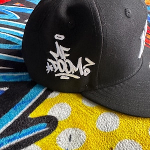 Peut inclure: Casquette de baseball noire avec texte blanc brodé de style graffiti sur le côté. La casquette repose sur une surface à motifs colorés. Le texte comprend les mots "VE DOOM".