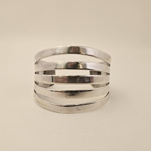 May include: A wide cuff bracelet crafted from polished silver-toned metal, featuring a multi-band design composed of five horizontal, curved bars that are connected at the sides to form a single, open-ended circular structure with a smooth finish.