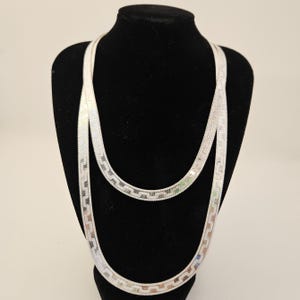 May include: A long, silver-toned herringbone chain necklace featuring a repeating geometric pattern along its flat, flexible surface. The necklace is draped in two loops, showcasing the metallic sheen and the intricate, interlocking link design.