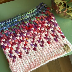 May include: A hand-knitted cowl in a chevron pattern. The cowl features a gradient of colors, starting with light pink at the bottom, transitioning to red, purple, blue, and green at the top. A small leather tag is attached.