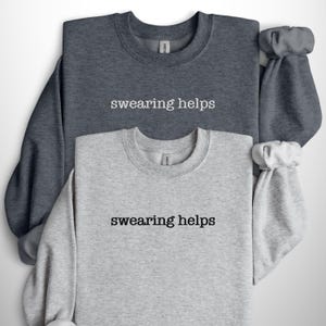 May include: Two crew neck sweatshirts, one dark gray and one light gray, are displayed. The phrase "swearing helps" is printed in black on the front of each sweatshirt. The sleeves are cuffed. The sweatshirts are made of a soft material.