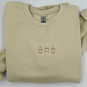 Può includere: Una felpa beige chiaro con tre tazze di caffè ricamate sul davanti. Le tazze sono marroni con coperchi bianchi e una cannuccia marrone.