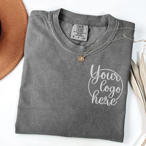 May include: A gray Comfort Colors t-shirt with the text "Your Logo Here" embroidered in white. A silver necklace with a small pendant is draped across the shirt. A brown hat is partially visible.