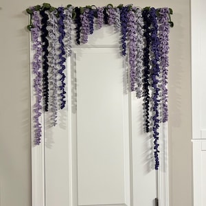 Peut inclure: Une guirlande de glycine au crochet, dans les tons de violet, lavande et vert, est suspendue au-dessus d'une porte blanche. La d&eacute;coration florale faite &agrave; la main pr&eacute;sente des brins de fleurs au crochet en cascade.