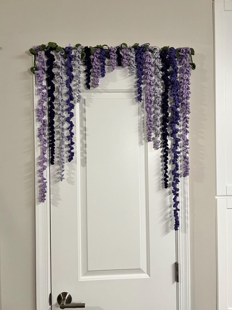 Peut inclure: Une guirlande de glycine au crochet, dans les tons de violet, lavande et vert, est suspendue au-dessus d'une porte blanche. La d&eacute;coration florale faite &agrave; la main pr&eacute;sente des brins de fleurs au crochet en cascade.