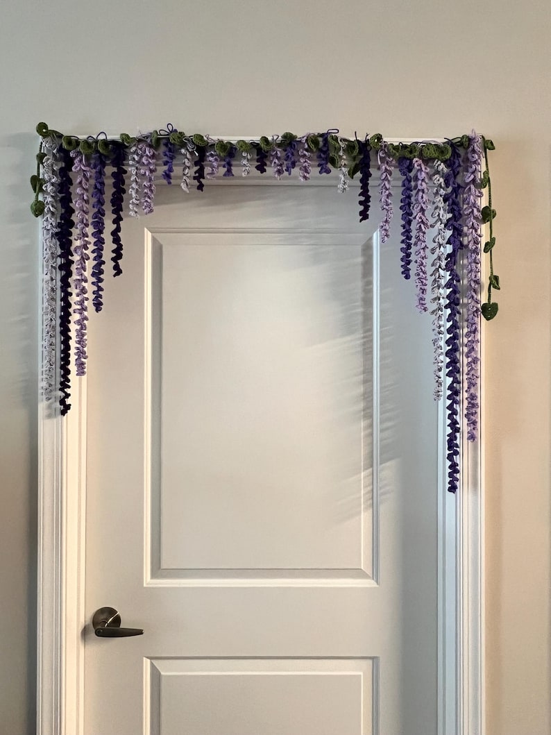 Peut inclure: Une guirlande de glycine au crochet, dans les tons de violet et de blanc, avec des feuilles vertes, est suspendue au-dessus d'une porte blanche. La guirlande est con&ccedil;ue pour imiter les fleurs de glycine en cascade.