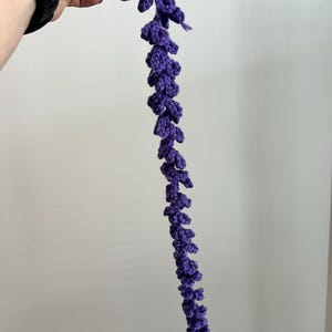 Peut inclure: Une tige florale violette au crochet faite &agrave; la main. La tige est d'un violet profond et pr&eacute;sente une s&eacute;rie de petits boutons floraux textur&eacute;s le long de sa longueur. La tige est tenue par une main sur un fond uni.