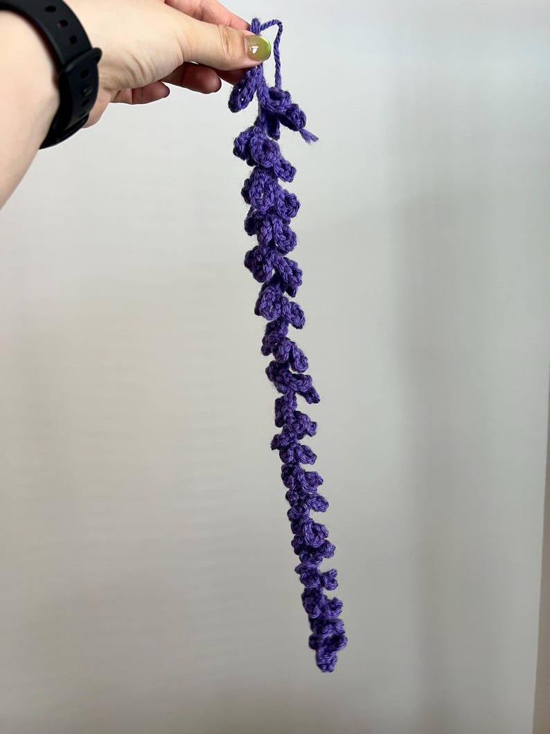 Peut inclure: Une tige florale violette au crochet faite &agrave; la main. La tige est d'un violet profond et pr&eacute;sente une s&eacute;rie de petits boutons floraux textur&eacute;s le long de sa longueur. La tige est tenue par une main sur un fond uni.
