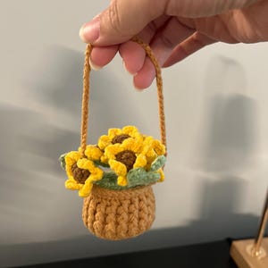 May include: A small, handmade crochet basket featuring a tan base and a matching handle. The basket is filled with three bright yellow sunflowers with brown centers and light green leaves, all crafted from yarn in a detailed, textured stitch.